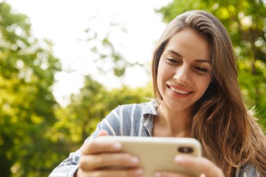 Parkta yürürken gülümseyen ve cep telefonu tutan Kafkasyalı esmer kadın.