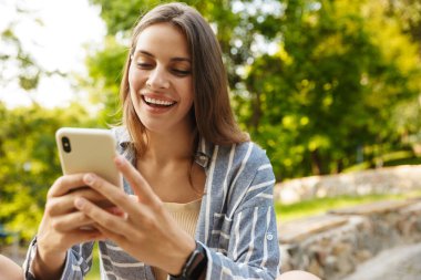 Parkta yürürken gülümseyen ve cep telefonu tutan Kafkasyalı esmer kadın.