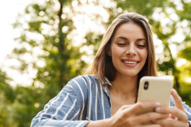 Parkta yürürken gülümseyen ve cep telefonu tutan Kafkasyalı esmer kadın.