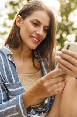 Parkta yürürken gülümseyen ve cep telefonu tutan Kafkasyalı esmer kadın.