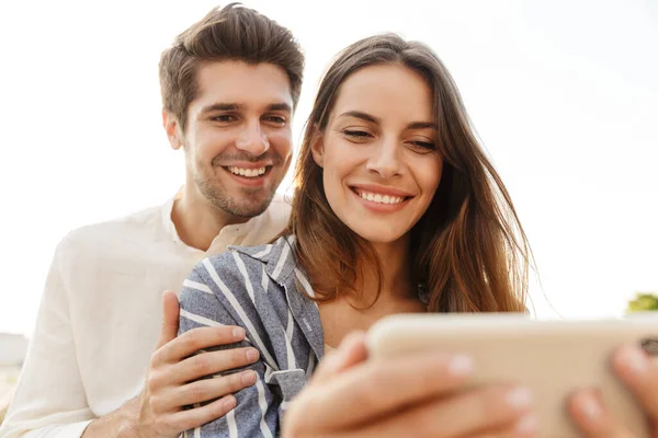 图片中年轻的高加索人快乐的夫妇坐在户外用手机时的微笑和拥抱 — 图库照片