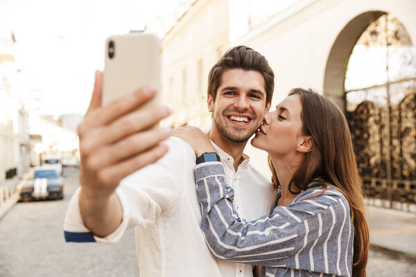 Image of young caucasian happy couple hugging and taking selfie photo while walking on city street