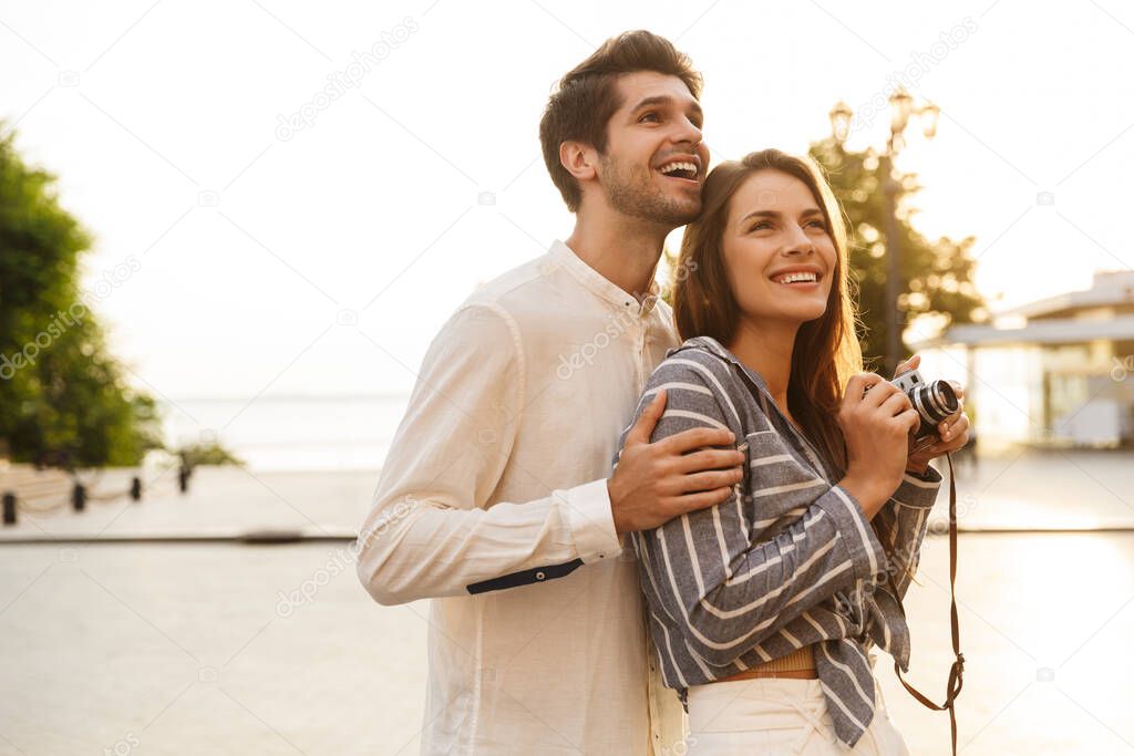 Image of young caucasian couple smiling and hugging while walking with retro camera through city street