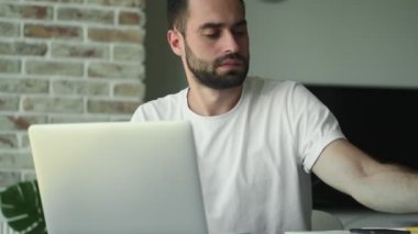 Evde dizüstü bilgisayar kullanan ve elinde kağıt tutan genç ciddi bir adam.
