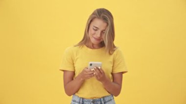 Sarı tişörtlü, mutlu, sarışın bir kadın sarı arka planda tek başına duran akıllı telefonunu kullanıyor.
