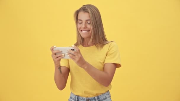 黄色のTシャツの若いブロンドの女性が遊んでいますゲームオン彼女のスマートフォンスタンディング孤立した上に黄色の背景 — ストック動画