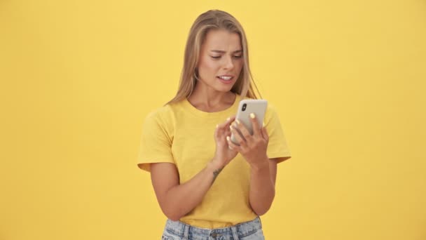 黄色の背景の上に隔離された彼女のスマートフォンを使用している間幸せな若い女性は何かを獲得しています — ストック動画