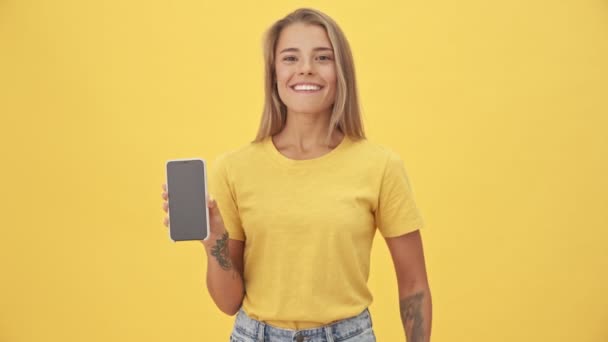 Uśmiechnięta Młoda Kobieta Wskazuje Ekran Swojego Smartfona Stojącego Żółtym Tle — Wideo stockowe