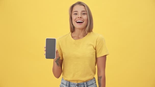 黄色の背景に隔離されたスマートフォンの画面を見せながら 喜んで若い女性が勝者のジェスチャーをしています — ストック動画