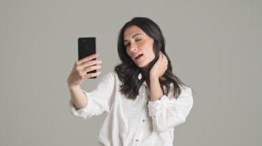 Çekici genç bir kadın, gri arka planda tek başına duran akıllı telefonunu kullanarak fotoğraf çekiyor.