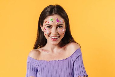 Yüzünde çıkartmalar olan, sarı arka planda izole edilmiş kameraya gülümseyen neşeli genç bir kadın resmi.