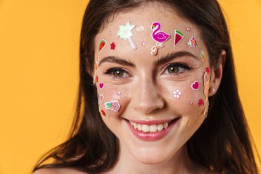 Yüzünde çıkartmalar olan, sarı arka planda izole edilmiş kameraya gülen neşeli genç bir kadın resmi.