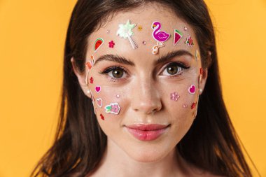 Yüzünde çıkartmalar olan genç bir kadının görüntüsü sarı arka planda izole edilmiş kameraya bakıyor.