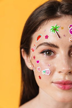 Yüzünde çıkartmalar olan, sarı arka planda izole edilmiş kameraya gülümseyen neşeli bir kadın görüntüsü.