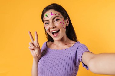 Sarı duvardan izole edilmiş selfie çekerken yüzünde çıkartmalar olan mutlu bir kadın resmi.