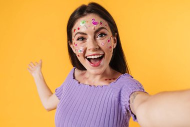 Yüzünde çıkartmalar olan mutlu bir kadın resmi. Kopyalama alanını gösterirken, sarı duvarda izole edilmiş selfie çekiyor.