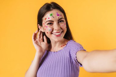 Sarı duvardan izole edilmiş selfie çekerken yüzünde çıkartmalar olan mutlu bir kadın resmi.