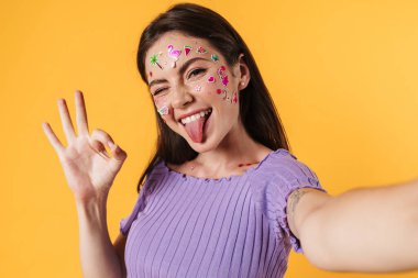 Sarı duvardan izole edilmiş selfie çekerken yüzünde çıkartmalar olan mutlu bir kadın resmi.