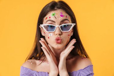 Yüzünde çıkartmalar olan, güneş gözlüğü takan neşeli bir kadın görüntüsü sarı arkaplanda öpüşen dudakları izole ediyor.