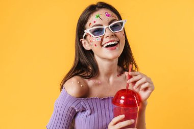 Yüzüne çıkartmalar yapıştırılmış, kokteyl içen ve sarı duvardan izole bir şekilde kahkahalar atan neşeli genç bir kadın.
