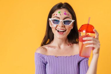 Yüzüne çıkartmalar yapıştırılmış, kokteyl içen ve sarı duvarda sırıtan neşeli genç bir kadın resmi.