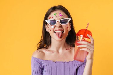 Yüzünde çıkartmalar olan, kokteyl içen ve sarı duvarda dilini gösteren neşeli bir kadın resmi.