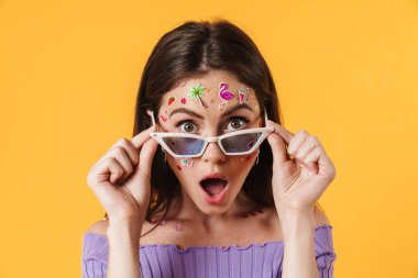 Suratı yapıştırılmış, güneş gözlüğü takmış, sarı duvarda izole edilmiş kameraya şaşırmış bir kadın görüntüsü.