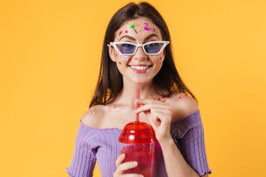 Yüzüne çıkartmalar yapıştırılmış, kokteyl içen ve sarı duvarda sırıtan neşeli genç bir kadın resmi.