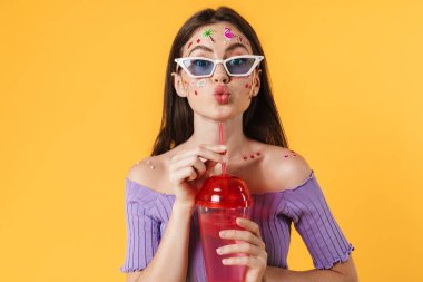 Yüzünde çıkartmalar olan, kokteyl içen ve sarı duvarda öpüşen dudakları olan neşeli genç bir kadın resmi.