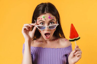 Yüzünde çıkartmalar olan, sarı duvarda izole edilmiş lolipop tutan genç bir kadın resmi.