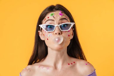 Yüzünde çıkartmalar olan komik bir kadın resmi. Sarı duvarın üzerinde sakız üflüyor.