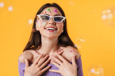 Yüzünde çıkartmalar olan, sarı arka planda izole edilmiş kameraya gülümseyen neşeli genç bir kadın resmi.