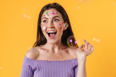 Yüzünde çıkartmalar olan, sarı arka planda izole edilmiş kameraya gülen genç ve heyecanlı bir kadın resmi.
