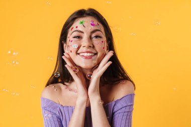 Yüzünde çıkartmalar olan, sarı arka planda izole edilmiş kameraya gülümseyen neşeli genç bir kadın resmi.