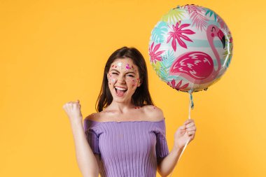 Yüzüne çıkartma yapıştırılmış, balon tutan ve sarı duvardan izole bir jest yapan heyecanlı bir kadın resmi.