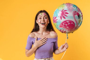 Yüzünde çıkartmalar olan, sarı arka planda izole edilmiş balon tutan heyecanlı genç bir kadın resmi