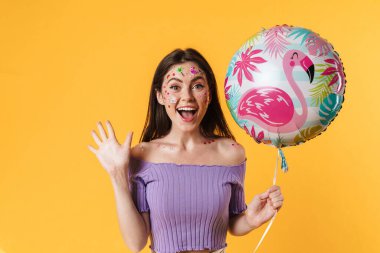 Yüzünde çıkartmalar olan, balon tutan ve sarı duvarda izole edilmiş bağıran heyecanlı bir kadın resmi.