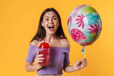Yüzünde çıkartmalar olan, balon tutan ve sarı duvarda izole edilmiş kokteyl içen bir kadın resmi.