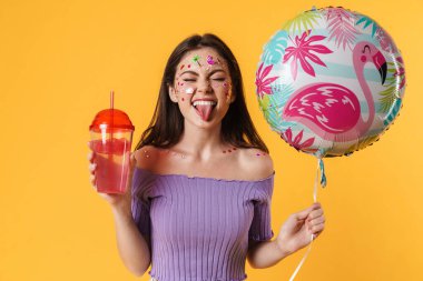 Yüzünde çıkartmalar olan, balon tutan ve sarı duvarda izole edilmiş kokteyl içen eğlenceli bir kadın resmi.