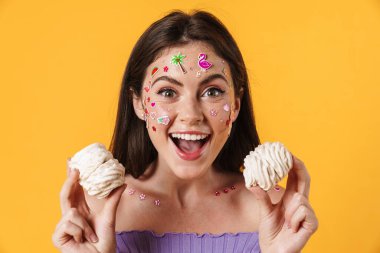 Yüzünde çıkartmalar olan, sarı arka planda izole edilmiş marşmelov tutan heyecanlı genç bir kadın resmi.