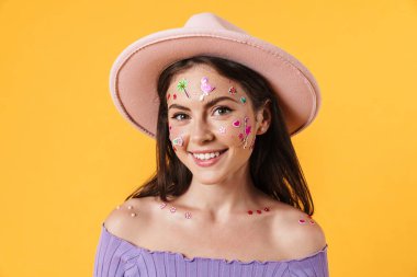 Yüzünde çıkartmalar olan, sarı arka planda izole edilmiş pembe şapkalı neşeli bir kadın resmi.