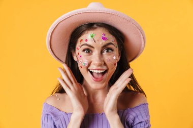 Yüzünde çıkartmalar olan, sarı arka planda izole edilmiş pembe şapkalı, heyecanlı genç bir kadın resmi