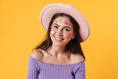 Yüzünde çıkartmalar olan, sarı arka planda izole edilmiş pembe şapkalı neşeli bir kadın resmi.