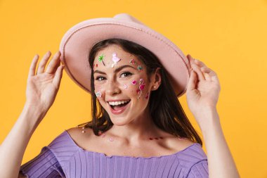 Yüzünde çıkartmalar olan, sarı arka planda izole edilmiş pembe şapkalı, gülen genç bir kadın resmi.