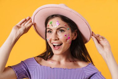 Yüzünde çıkartmalar olan, sarı arka planda izole edilmiş pembe şapkalı, gülen genç bir kadın resmi.