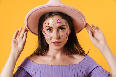 Yüzünde çıkartmalar olan, sarı arka planda izole edilmiş pembe şapkalı çekici bir kadın resmi.