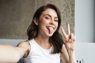 Evde selfie çekerken dilini ve barış işaretini gösteren güzel, neşeli bir kadın görüntüsü