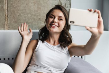 Neşeli bir kadının el sallaması ve evdeki kanepede dinlenirken akıllı telefondan selfie çekmesi.