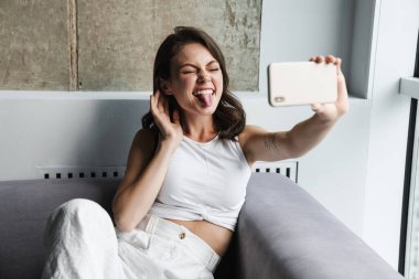 Neşeli bir kadın görüntüsü. Dilini gösteriyor ve evinde kanepede dinlenirken akıllı telefondan selfie çekiyor.
