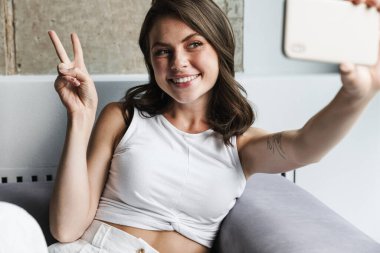 Neşeli bir kadının huzur jesti yapması ve evdeki kanepede dinlenirken akıllı telefonuyla selfie çekmesi.